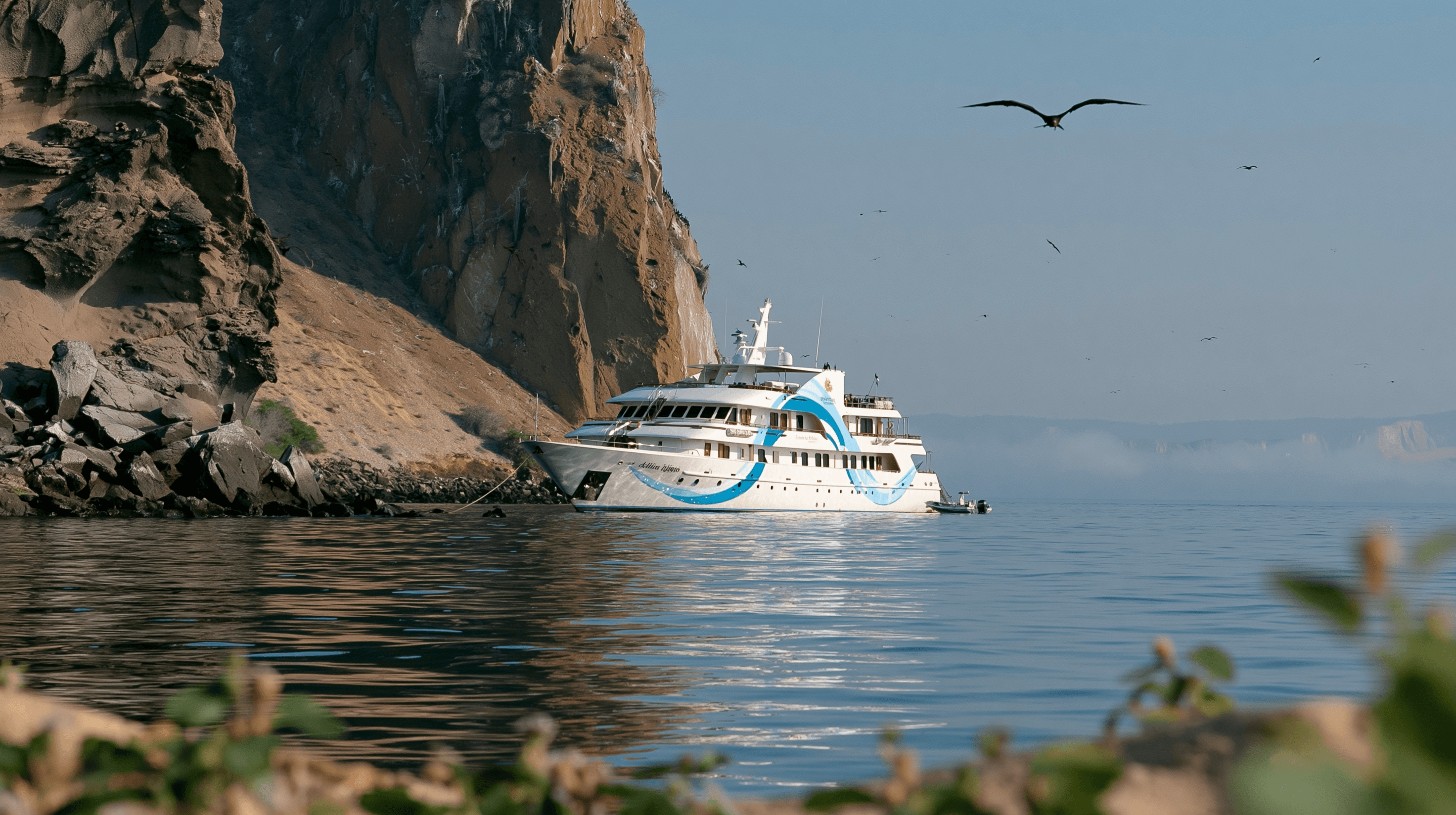 A small Galápagos expedition ship at anchor under a sea-cliff with frigatebirds overhead at first light.